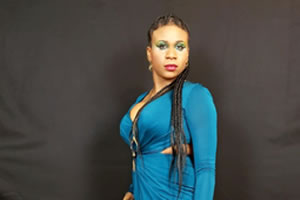 Lachi, Founder. A black woman with cornrows and green makeup wearing an aqua marine dress.