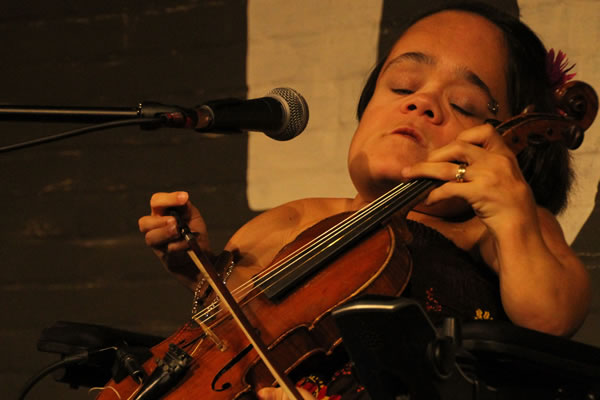 Gaelynn Lea, Co-Founder. A white woman with a flower in her hair, holding a violin in front of a mic.