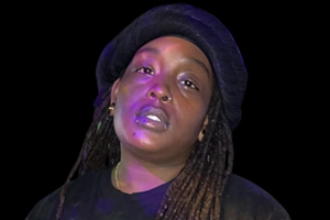 Jai Yoko, Secretary. A Black woman wearing a slouchy black hat and black shirt.