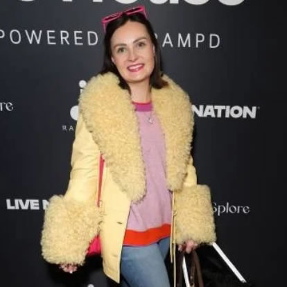 a woman with dark hair, a fluffy butter yellow coat and a cane in one hand stands in front of a banner for Rampd's DisCo house event