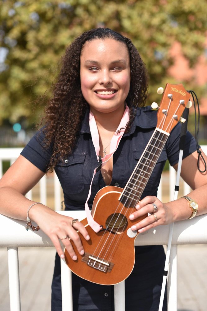 A Latina with dark, curly hair smiling and holding on to her ukulele while standing outside.