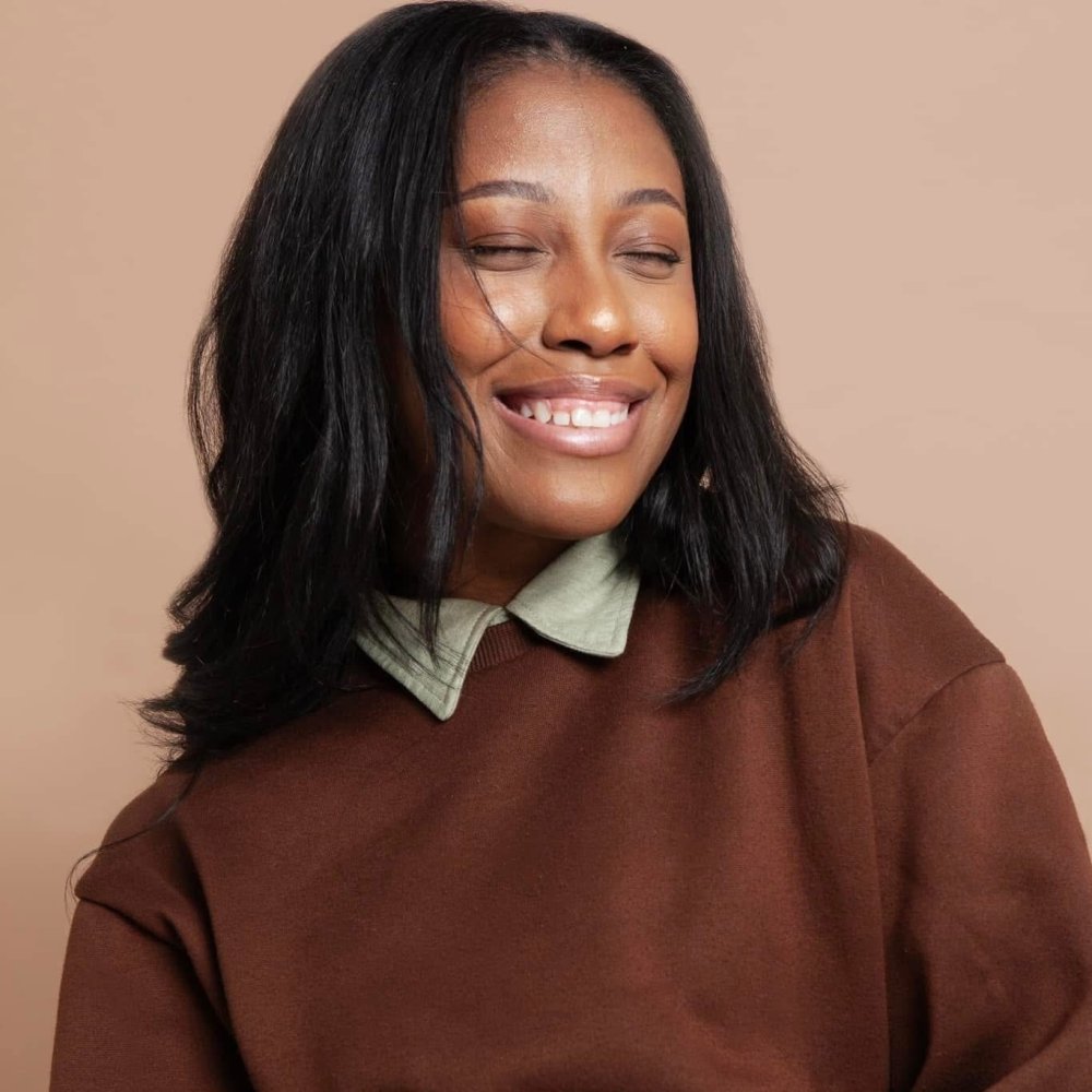 Indi smiles with shoulder-length black hair wearing a brown sweater over a light-colored collared shirt.