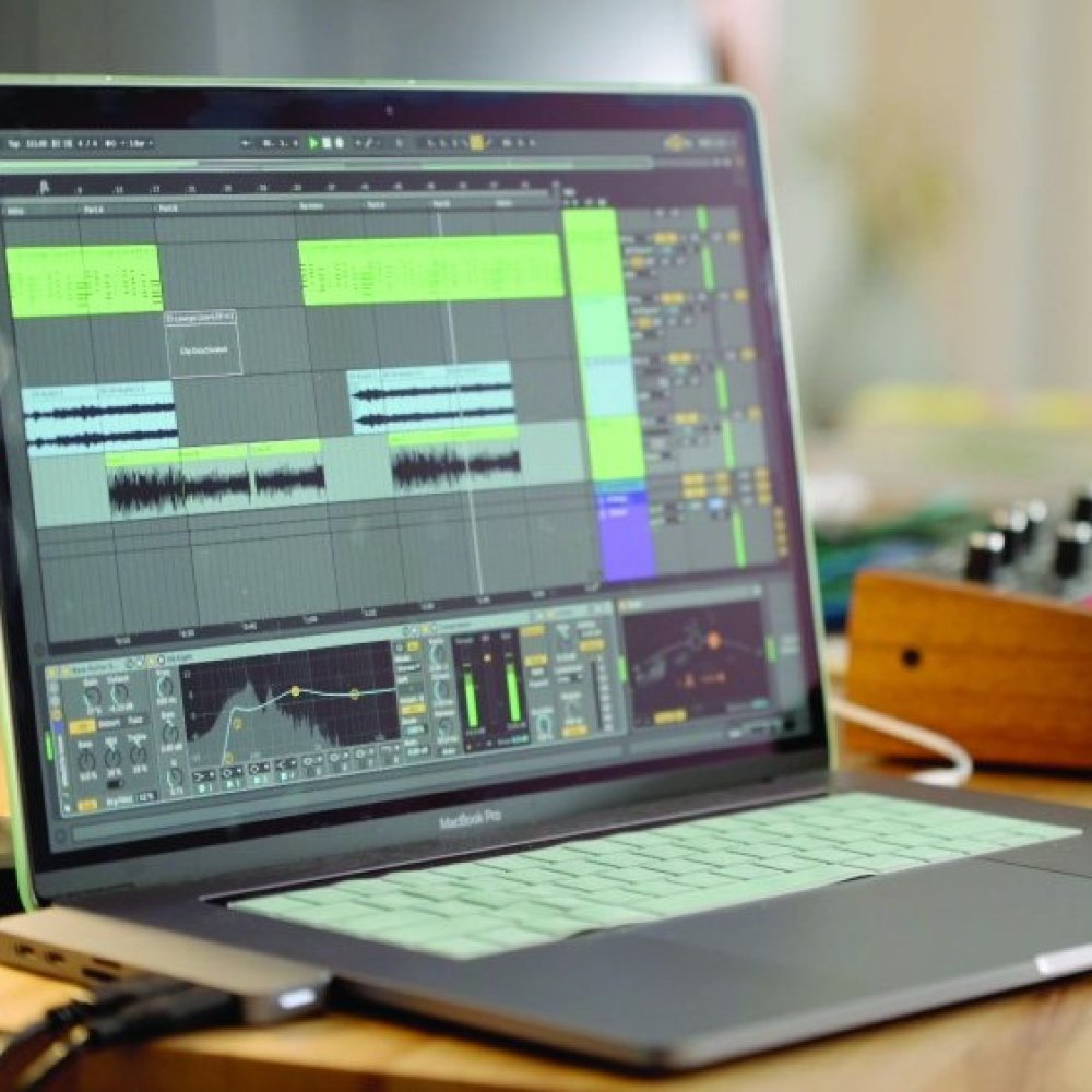 An opened Macbook pro laptop with Ableton opened on the screen on a desk next to a midi controller.