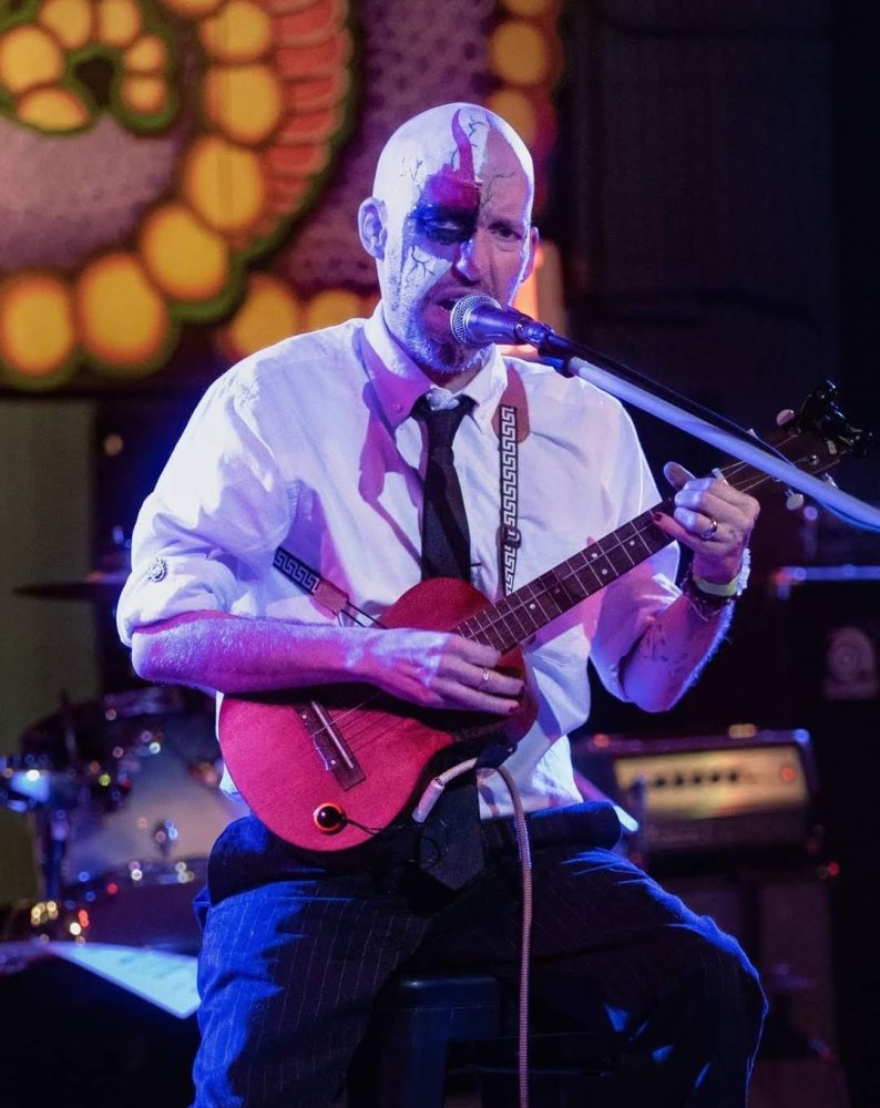 Garrett DeVaughn, the Mad Uke-Bard.  Sitting on a bar stool with black pants, white shirt, black tie.  Playing metal and rock on a Tenor Ukulele.  Right side of face/head in body paint.