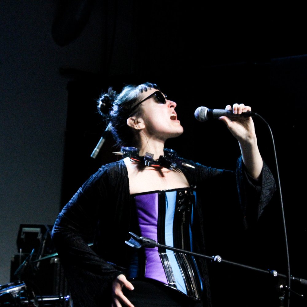 A tarot-card-style portrait of Tori / DEĀ with a black reflective blindness cane, black hair, glasses, piercings, and a trench coat, wearing a crown, with a hopeful look on their face. The card, labeled “The Ascension,” features alchemical symbols depicting water, a serpent, and different goddesses. The card is drawn by US Game Systems tarot card artist Sabina Espinet.