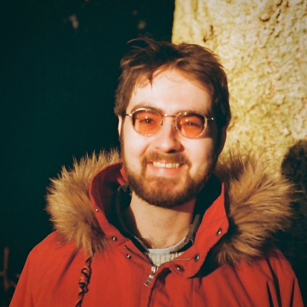 Jay Pocknell wearing a red parka jacket in front of a tree at sunset.