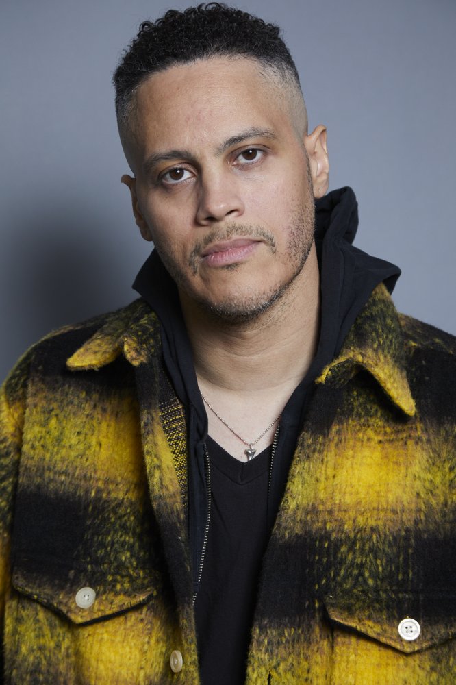 James Ian - A Black man with short hair wearing a yellow and black flannel shirt. He wears and a black hoodie and black t-shirt underneath the flannel.