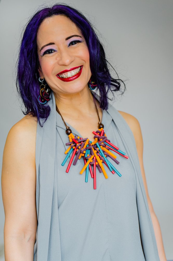 Lucy is smiling broadly, with dark purple hair in the front part, and a colorful wooden necklace on.